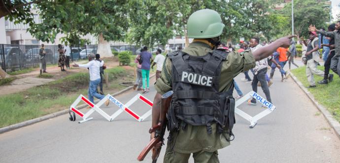 Un homme habillé en uniforme de police se tient à l'avant plan de l'image, dos à la caméra. Des manifestant·e·s sont dispersé·e·s à l'arrière-plan.