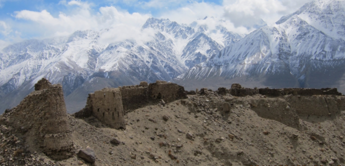 L'image montre un paysage accidenté et époustouflant d'un village pamiri niché dans les montagnes du Pamir, au Tadjikistan. Les ruines d'anciennes structures en pierre sont visibles au premier plan, perchées sur un terrain sec et rocailleux. À l'arrière-plan, les imposants pics enneigés de la chaîne du Pamir s'élèvent de façon spectaculaire dans un ciel bleu partiellement nuageux.