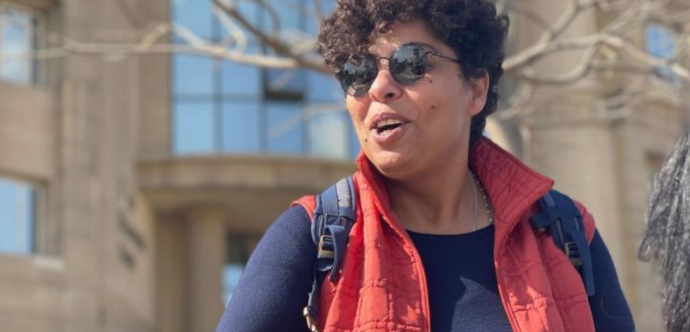 Photo de Rasha Azab en exterieur. Elle porte des lunettes rondes noires et un sans manche rouge.