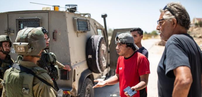 Un groupe d’hommes discute avec des soldats équipés de casques et d’armes, près d’un véhicule blindé militaire. Un homme en t-shirt rouge et casquette, tenant un téléphone, semble gesticuler en parlant. La scène se déroule sur une route en plein soleil, avec quelques bâtiments visibles au loin.