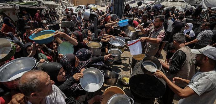 Des bénévoles distribuent de la nourriture à une foule dense de personnes, femmes, hommes et enfants, tendant des casseroles, bols et seaux dans une zone urbaine détruite. L’image reflète une situation d’urgence humanitaire, avec des bâtiments en ruine et des signes de conflit à l’arrière-plan.