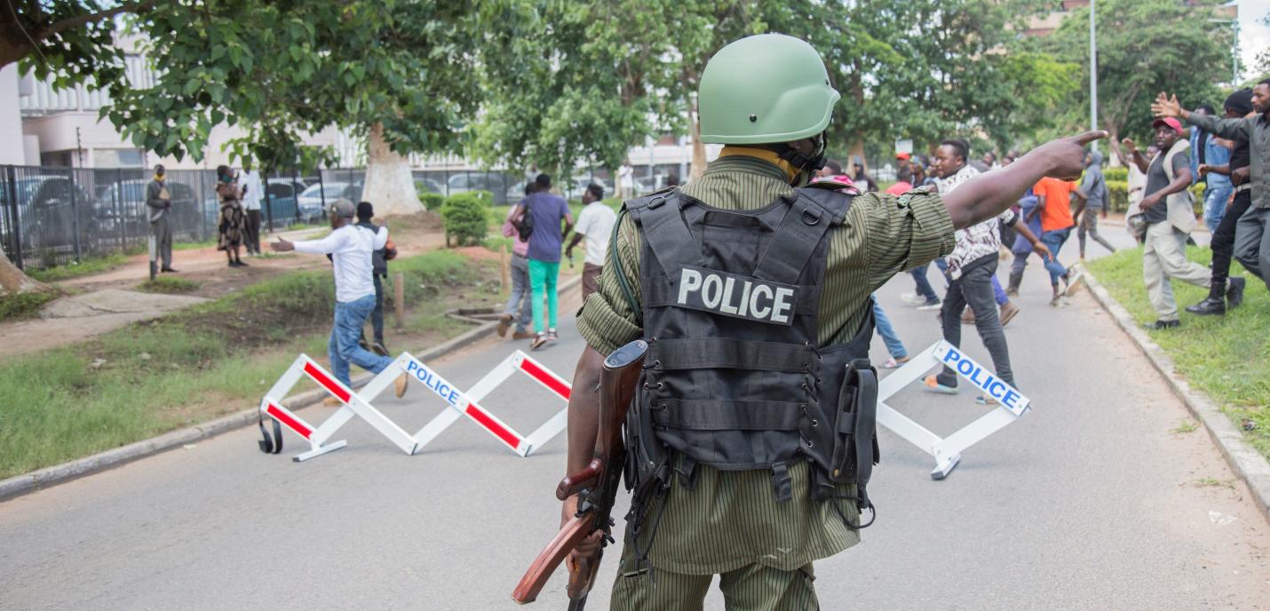 Un homme habillé en uniforme de police se tient à l'avant plan de l'image, dos à la caméra. Des manifestant·e·s sont dispersé·e·s à l'arrière-plan.