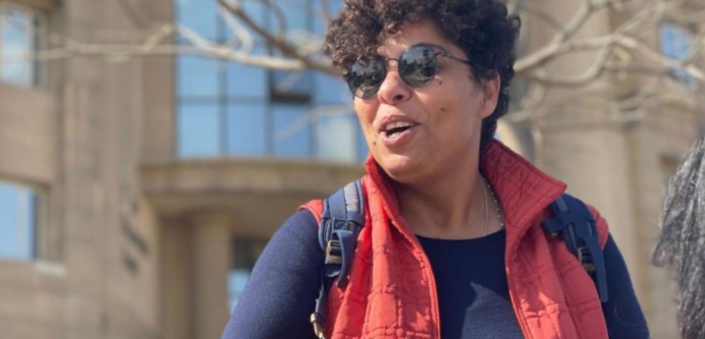 Photo de Rasha Azab en exterieur. Elle porte des lunettes rondes noires et un sans manche rouge.