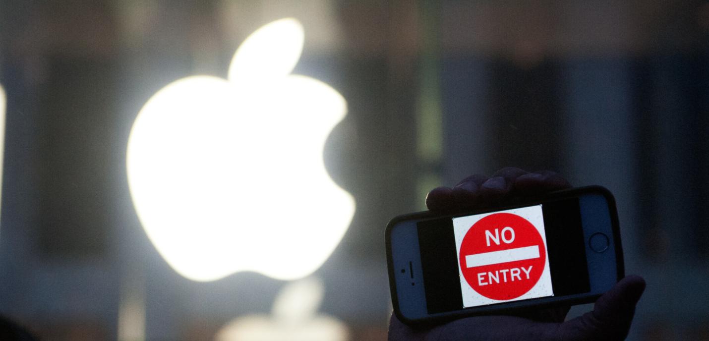 Un téléphone tenu dans une main affiche le message "No Entry" sur son écran, tandis qu'en arrière-plan, le logo lumineux d'un magasin Apple.