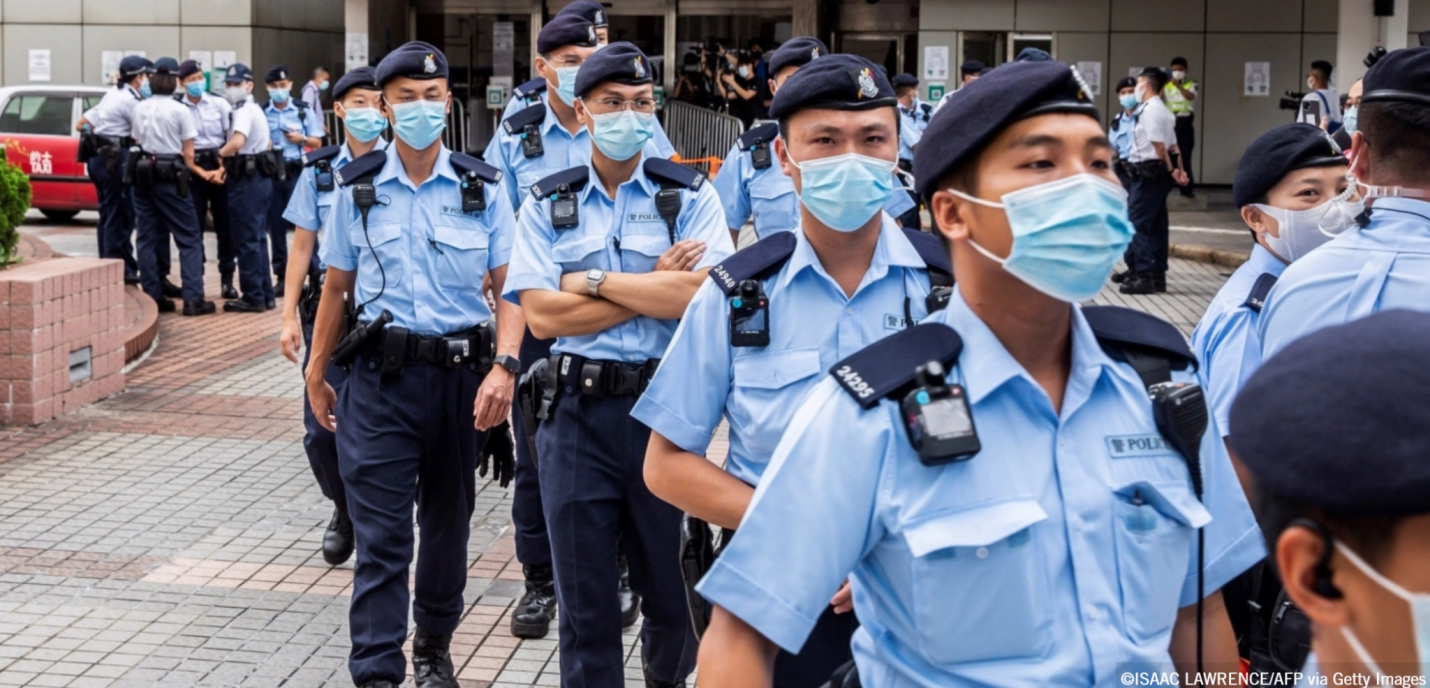 Groupe de policiers en uniforme bleu clair, portant des masques chirurgicaux et des bérets, marchant en file dans un espace urbain pavé. Certains portent des caméras corporelles et des équipements à la ceinture. À l’arrière-plan, d’autres agents discutent entre eux près d’un bâtiment officiel.