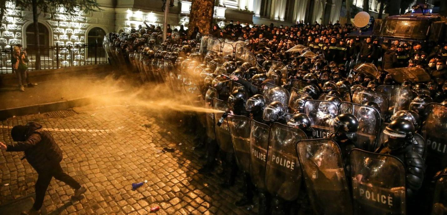 Un groupe compact de policiers antiémeute, protégés par des boucliers transparents et casques, fait face à des manifestants de nuit. Un policier utilise un gaz irritant ou spray poivre contre un manifestant replié en avant. La scène se déroule sur une rue pavée, dans une ambiance tendue et violente, avec une forte présence policière en arrière-plan.