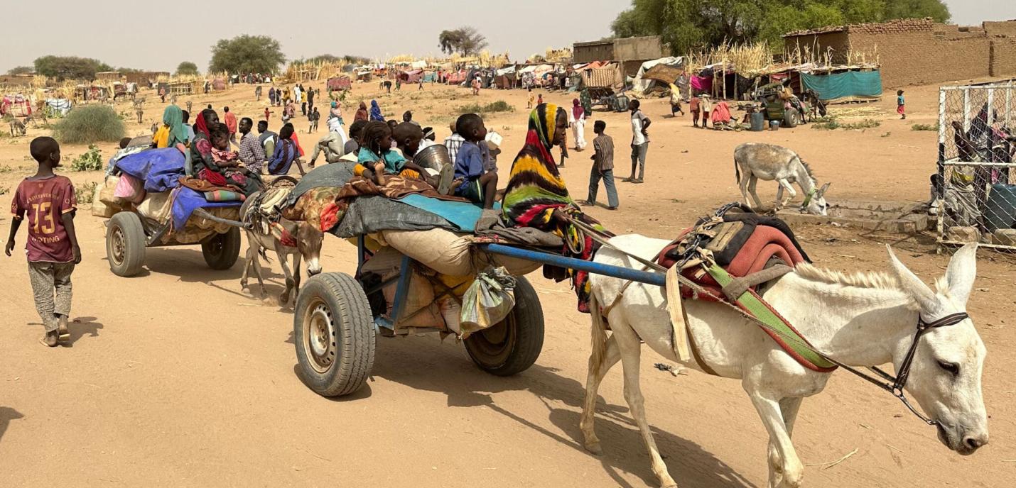 Scène de vie dans un camp de réfugiés en milieu désertique. Des ânes tirent des charrettes chargées de sacs, d'effets personnels et de plusieurs enfants assis, certains enveloppés dans des couvertures colorées. Des adultes marchent à côté, et en arrière-plan, on voit de nombreuses personnes, des abris de fortune, des huttes et des arbres. Le sol est sableux, et l'atmosphère suggère chaleur et aridité.