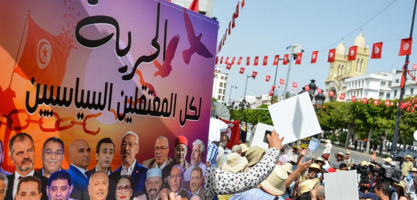 « Manifestation en Tunisie avec des drapeaux tunisiens suspendus en arrière-plan et une foule brandissant des pancartes. Au premier plan, une grande affiche en arabe avec des portraits de personnalités politiques et le mot "Liberté" en gros caractères, demandant la libération des prisonniers politiques. La scène se déroule en plein air, près d’un édifice historique visible au fond.