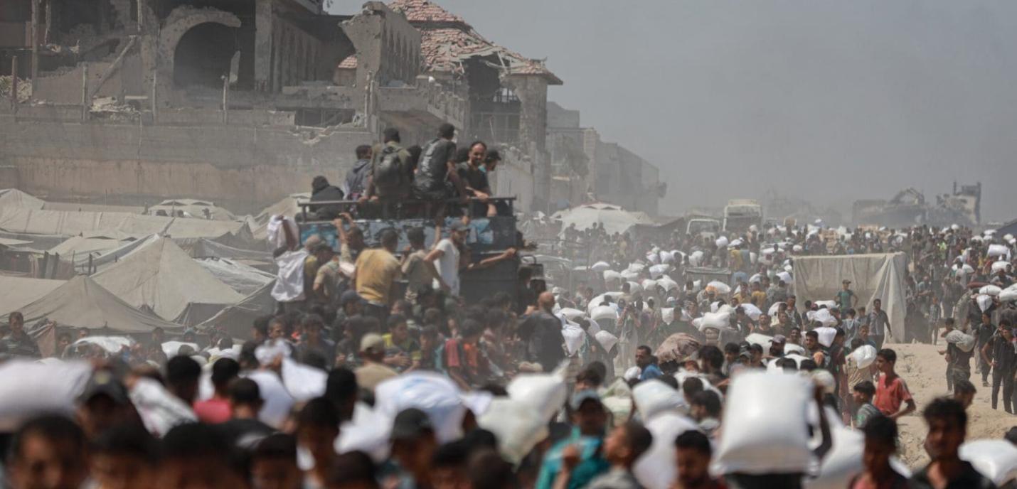 Une foule dense de personnes marche dans une zone poussiéreuse et dévastée, portant des sacs blancs sur leurs épaules, probablement de l'aide humanitaire. Des tentes et des bâtiments en ruine apparaissent à l'arrière-plan, témoignant d'un contexte de conflit ou de crise humanitaire majeure. Des véhicules et des soldats sont visibles au milieu de la scène, accentuant l'atmosphère de déplacement de masse et de survie.