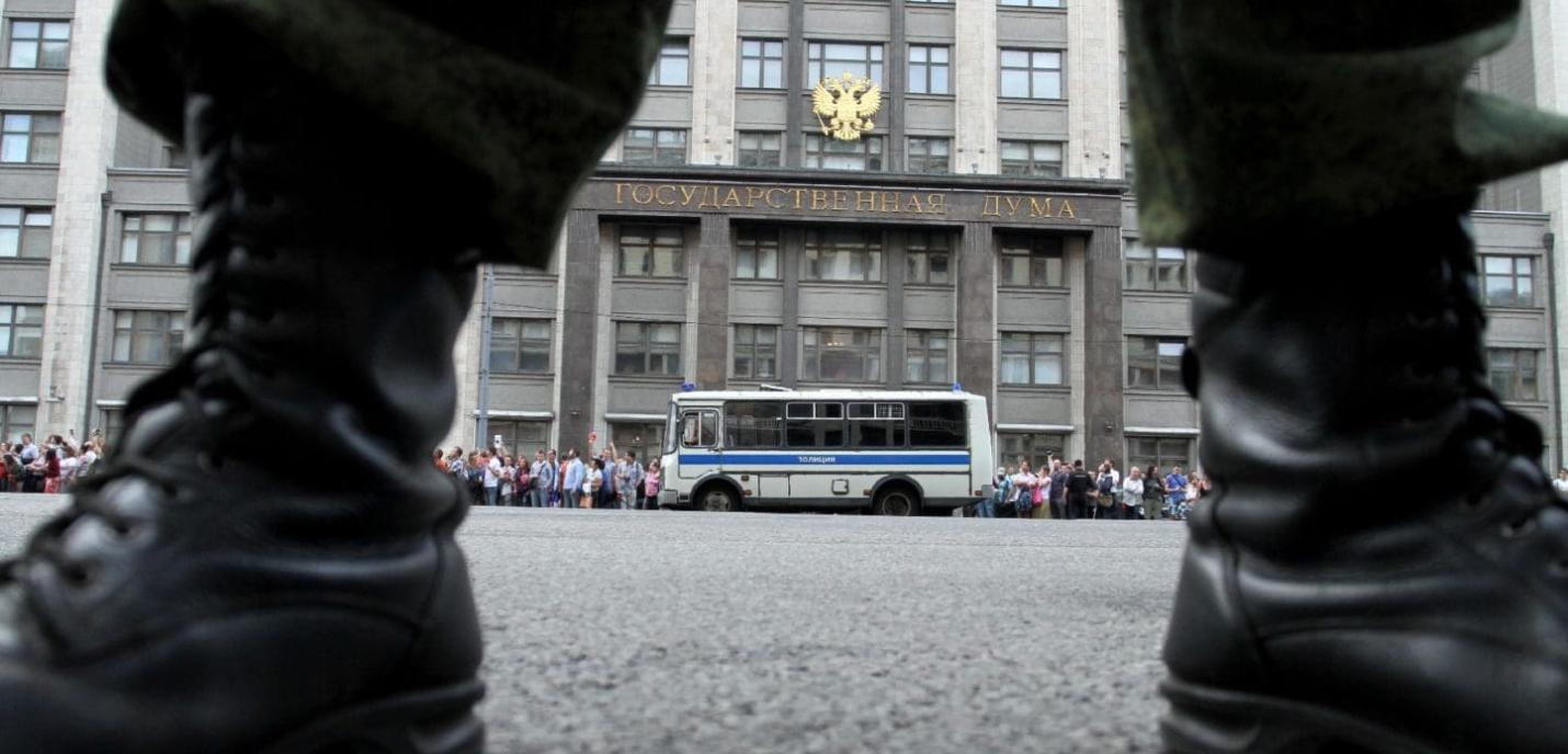 Vue au ras du sol entre deux bottes de militaire ou de policier, face au bâtiment de la Douma d’État en Russie. Une foule de personnes se tient en file le long du trottoir, à côté d’un bus de police stationné. La scène suggère une manifestation ou un rassemblement surveillé devant une institution gouvernementale.