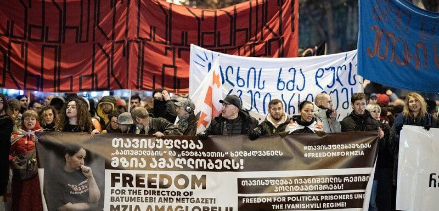 Manifestation de nuit avec une foule brandissant des banderoles. Au premier plan, une large bannière demande la liberté pour Mzia Amaglobeli, directrice de Batumelebi et Netgazeti. D'autres bannières contiennent des messages en géorgien et en anglais, appelant à la libération des prisonniers politiques