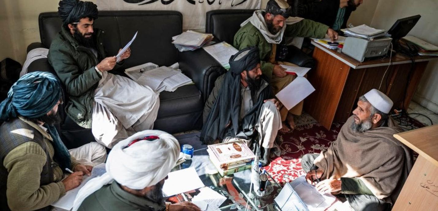 Un groupe d’hommes barbus portant des turbans et des vêtements traditionnels afghans est réuni dans une petite pièce. Certains sont assis sur un canapé ou au sol, d’autres travaillent à un bureau avec un ordinateur et des piles de documents. Ils semblent discuter et examiner des papiers. En arrière-plan, une grande bannière blanche ornée d’inscriptions en arabe est accrochée au mur.