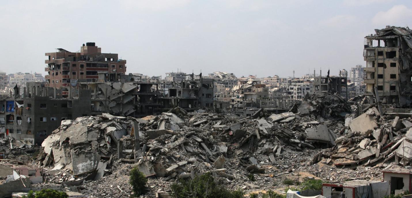 Vue panoramique d’une ville fortement détruite, avec des immeubles effondrés et des tas de gravats. Au premier plan, quelques arbres et petites structures encore debout contrastent avec les ruines massives en arrière-plan. Le ciel est nuageux, accentuant l’atmosphère de désolation