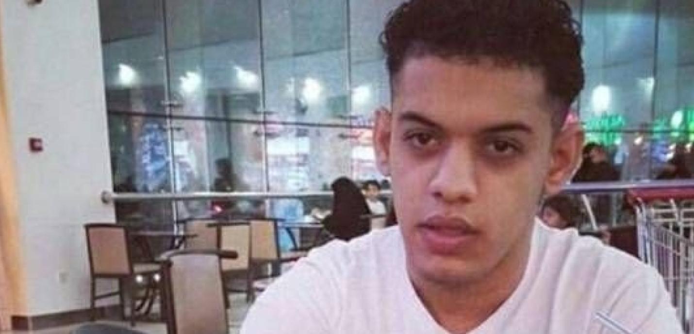 Un jeune homme assis dans un centre commercial, portant un t-shirt blanc, regarde droit vers la caméra. En arrière-plan, on distingue des tables, des chaises et des lumières de magasins à travers de grandes baies vitrées.
