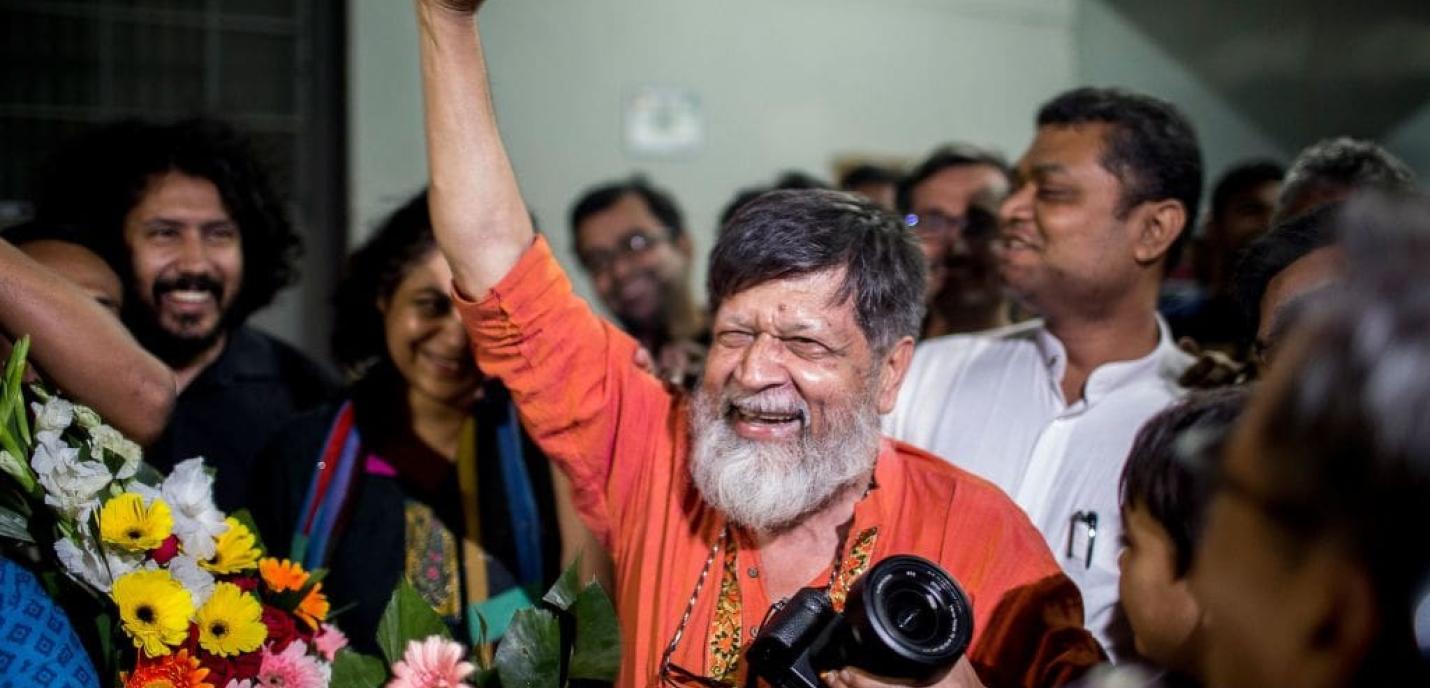 Un homme âgé, souriant largement, lève le poing en signe de victoire au milieu d’une foule joyeuse. Il porte une chemise orange et tient un appareil photo. Des bouquets de fleurs colorées sont visibles au premier plan, tandis que les personnes autour de lui applaudissent et sourient