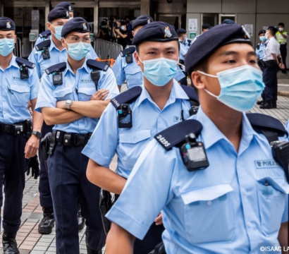 Groupe de policiers en uniforme bleu clair, portant des masques chirurgicaux et des bérets, marchant en file dans un espace urbain pavé. Certains portent des caméras corporelles et des équipements à la ceinture. À l’arrière-plan, d’autres agents discutent entre eux près d’un bâtiment officiel. 