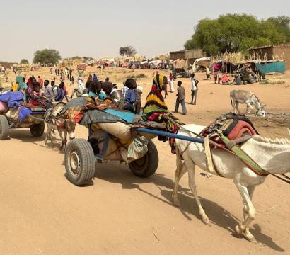 Scène de vie dans un camp de réfugiés en milieu désertique. Des ânes tirent des charrettes chargées de sacs, d'effets personnels et de plusieurs enfants assis, certains enveloppés dans des couvertures colorées. Des adultes marchent à côté, et en arrière-plan, on voit de nombreuses personnes, des abris de fortune, des huttes et des arbres. Le sol est sableux, et l'atmosphère suggère chaleur et aridité. 