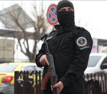 « Un homme en uniforme noir, masqué et tenant un fusil d’assaut, se tient en position de garde dans une rue. À l’arrière-plan, un piéton en capuche marche devant des voitures, dont un taxi jaune, et des panneaux urbains, par temps couvert. 