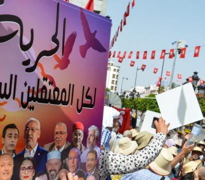 « Manifestation en Tunisie avec des drapeaux tunisiens suspendus en arrière-plan et une foule brandissant des pancartes. Au premier plan, une grande affiche en arabe avec des portraits de personnalités politiques et le mot "Liberté" en gros caractères, demandant la libération des prisonniers politiques. La scène se déroule en plein air, près d’un édifice historique visible au fond.
