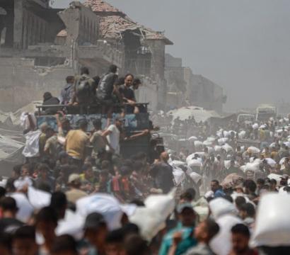 Une foule dense de personnes marche dans une zone poussiéreuse et dévastée, portant des sacs blancs sur leurs épaules, probablement de l'aide humanitaire. Des tentes et des bâtiments en ruine apparaissent à l'arrière-plan, témoignant d'un contexte de conflit ou de crise humanitaire majeure. Des véhicules et des soldats sont visibles au milieu de la scène, accentuant l'atmosphère de déplacement de masse et de survie. 