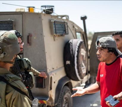 Un groupe d’hommes discute avec des soldats équipés de casques et d’armes, près d’un véhicule blindé militaire. Un homme en t-shirt rouge et casquette, tenant un téléphone, semble gesticuler en parlant. La scène se déroule sur une route en plein soleil, avec quelques bâtiments visibles au loin.