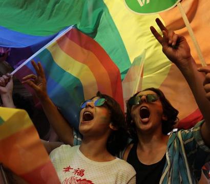 Deux personnes portant des lunettes de soleil et criant de joie sous un grand drapeau arc-en-ciel lors d'une manifestation pour les droits LGBTQIA+. Elles lèvent les bras en signe de victoire. L'ambiance est festive et militante, entourées de drapeaux multicolores.