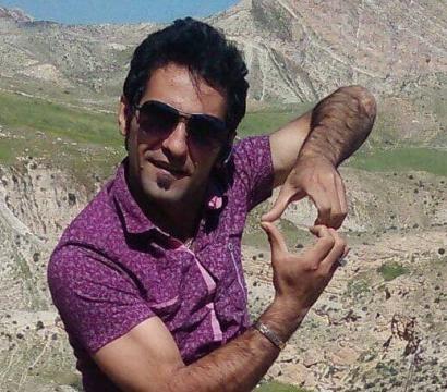 « Un homme portant une chemise violette à motifs et des lunettes de soleil pose en plein air, devant un paysage montagneux aride et vallonné. Il forme un geste avec ses mains comme s’il encadrait quelque chose entre ses doigts, en souriant légèrement à la caméra. 