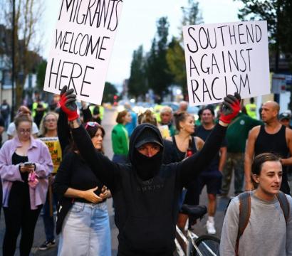 Manifestation dans une rue avec un groupe de personnes. Au centre, une personne masquée vêtue de noir tient deux pancartes en l'air indiquant : "Migrants welcome here" et "Southend against racism". D'autres manifestants sont visibles en arrière-plan