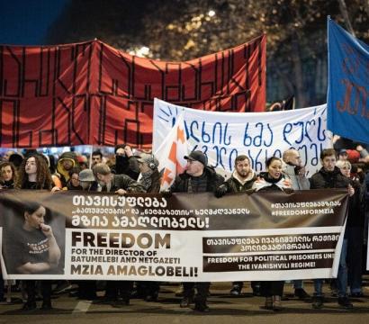Manifestation de nuit avec une foule brandissant des banderoles. Au premier plan, une large bannière demande la liberté pour Mzia Amaglobeli, directrice de Batumelebi et Netgazeti. D'autres bannières contiennent des messages en géorgien et en anglais, appelant à la libération des prisonniers politiques