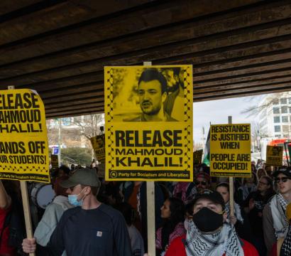 Manifestation sous un pont avec des pancartes jaunes portant des slogans en anglais tels que « Release Mahmoud Khalil ! », « Hands off our students ! » et « Resistance is justified when people are occupied ! ». Une foule de personnes, certaines portant un keffieh ou un masque, brandit des pancartes et un drapeau palestinien, exprimant leur solidarité avec la Palestine.