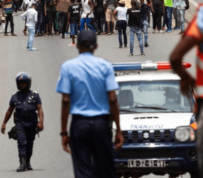 « Manifestation sur une route, avec un groupe de personnes rassemblées en arrière-plan. Au premier plan, plusieurs policiers, certains portant des casques et des gilets, encadrent la scène, tandis qu'une voiture de police marquée "Trânsito" est visible. Un policier en gilet fluorescent observe la situation