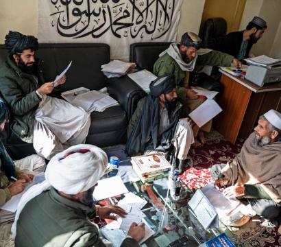 Un groupe d’hommes barbus portant des turbans et des vêtements traditionnels afghans est réuni dans une petite pièce. Certains sont assis sur un canapé ou au sol, d’autres travaillent à un bureau avec un ordinateur et des piles de documents. Ils semblent discuter et examiner des papiers. En arrière-plan, une grande bannière blanche ornée d’inscriptions en arabe est accrochée au mur.