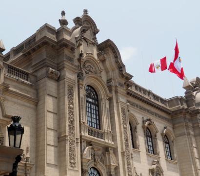 Un bâtiment gouvernemental imposant de style colonial, avec une façade en pierre richement décorée et de grandes colonnes. Deux drapeaux du Pérou flottent au sommet du bâtiment, sur fond de ciel bleu clair.