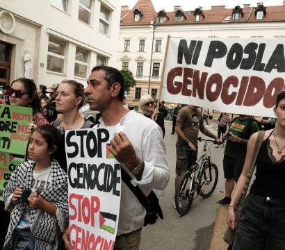 Un groupe de personnes participe à une manifestation dans la rue. Elles tiennent des pancartes portant des slogans contre le génocide et en soutien à Gaza. On peut lire « STOP GENOCIDE », « GAZA CAN'T WAIT » et « NI POSLA Z GENOCIDOM ». Les manifestants, dont des adultes et un enfant portant un keffieh, affichent des expressions sérieuses. L’ambiance est calme mais déterminée.