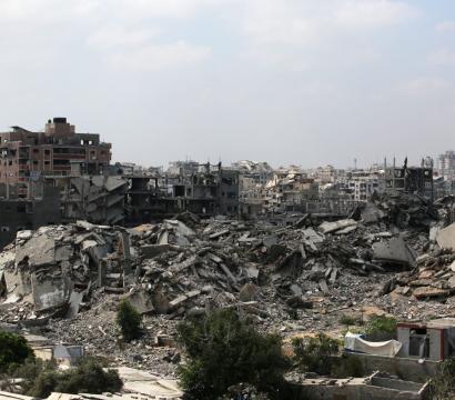 Vue panoramique d’une ville fortement détruite, avec des immeubles effondrés et des tas de gravats. Au premier plan, quelques arbres et petites structures encore debout contrastent avec les ruines massives en arrière-plan. Le ciel est nuageux, accentuant l’atmosphère de désolation