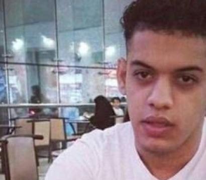 Un jeune homme assis dans un centre commercial, portant un t-shirt blanc, regarde droit vers la caméra. En arrière-plan, on distingue des tables, des chaises et des lumières de magasins à travers de grandes baies vitrées.