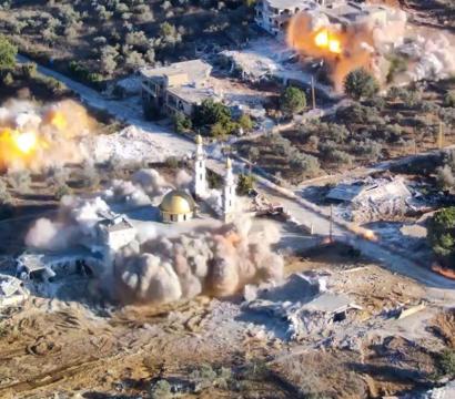 Vue aérienne d’un village touché par des bombardements, avec plusieurs explosions visibles et un épais nuage de fumée s’élevant autour d’une mosquée à coupole dorée, au milieu de bâtiments détruits et de zones rurales.