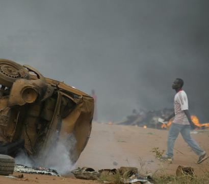 Un véhicule retourné et brûlé, encore fumant, repose au milieu d’une rue poussiéreuse. À l’arrière-plan, une épaisse fumée noire s’élève dans le ciel, avec des flammes visibles près de débris éparpillés. Un homme vêtu d’un t-shirt blanc à manches courtes avec des rayures rouges et d’un jean marche calmement à proximité, semblant ignorer la scène chaotique autour de lui. L’atmosphère évoque des troubles civils ou des affrontements récents.