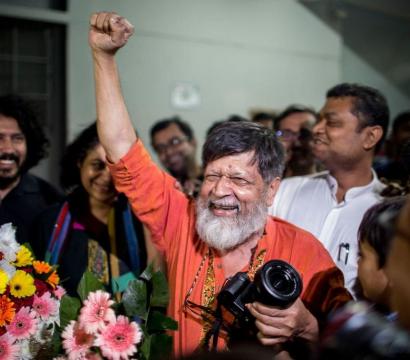 Un homme âgé, souriant largement, lève le poing en signe de victoire au milieu d’une foule joyeuse. Il porte une chemise orange et tient un appareil photo. Des bouquets de fleurs colorées sont visibles au premier plan, tandis que les personnes autour de lui applaudissent et sourient