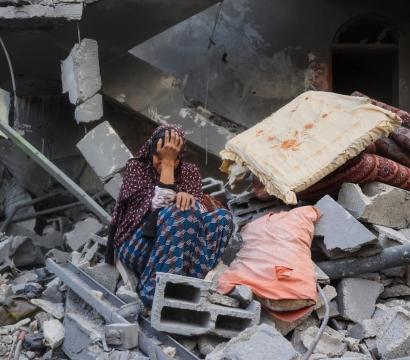 Une femme assise au milieu des décombres d’un bâtiment détruit, la tête dans la main en signe de détresse. Autour d’elle, des blocs de béton brisés, des poutres métalliques tordues et quelques matelas recouverts de poussière témoignent de la destruction causée par le bombardement.
